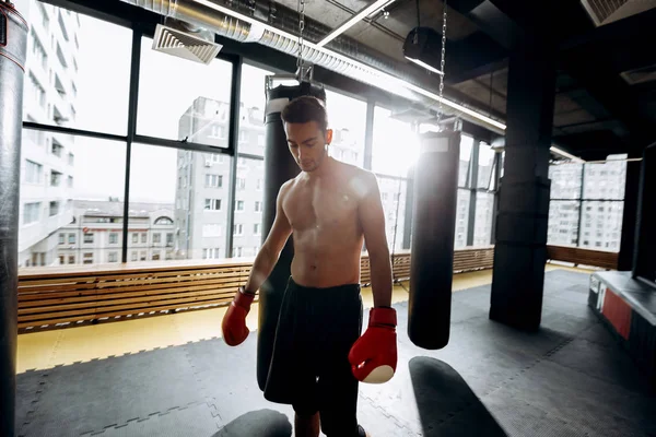 Sportsman en gants de boxe rouges avec un torse nu habillé dans le short noir se tient à côté des sacs de boxe dans la salle de gym — Photo