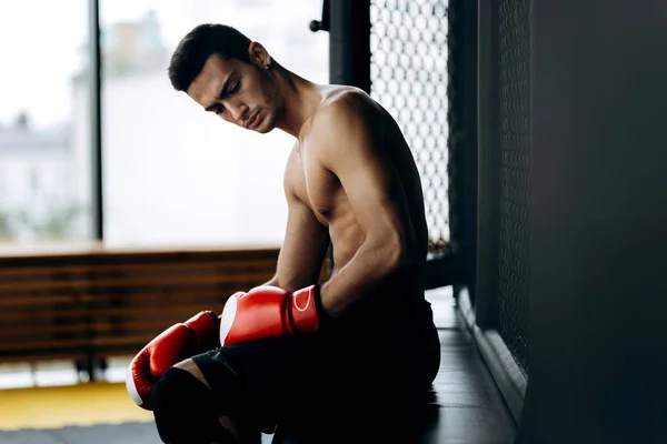 Brutal cara de cabelos escuros com um tronco nu e as luvas de boxe vermelho em suas mãos senta-se na borda do anel de boxe — Fotografia de Stock