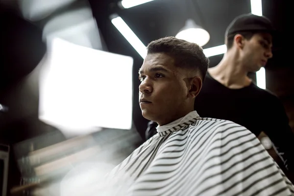 Mode Barberaren i svarta kläder gör en rakhyvel klippa hår för en eleganta svarthåriga mannen sitter i fåtöljen i en snygg barbershop — Stockfoto