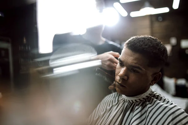 Mode Barberaren i svarta kläder gör en rakhyvel klippa hår för en eleganta svarthåriga mannen sitter i fåtöljen i en snygg barbershop — Stockfoto