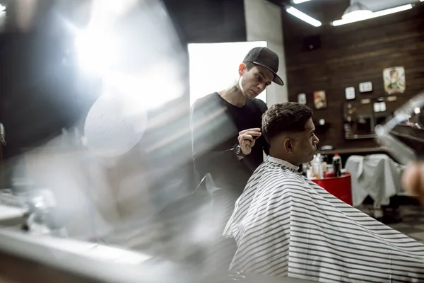 The fashion barber in black clothes makes a razor cut hair for a black-haired man sitting in the armchair in a barbershop with lots of light