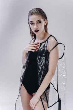 Genç kız koyu ruj ile ince ve ıslak saç yüzme takım elbise ve şeffaf yağmur ceket siyah giyimli stüdyoda beyaz zemin üzerine poz