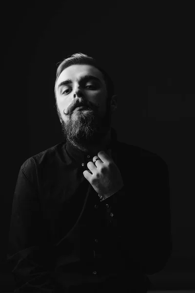 Retrato preto e branco de um homem elegante com barba e penteado elegante vestido com a camisa preta no fundo preto — Fotografia de Stock