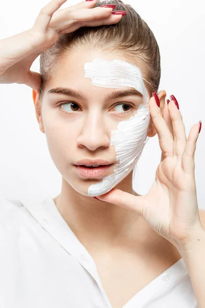 Young woman dressed in white clothes with a white cosmetic mask on a half of her face holds her hands at her face on the white background — Stock Photo, Image