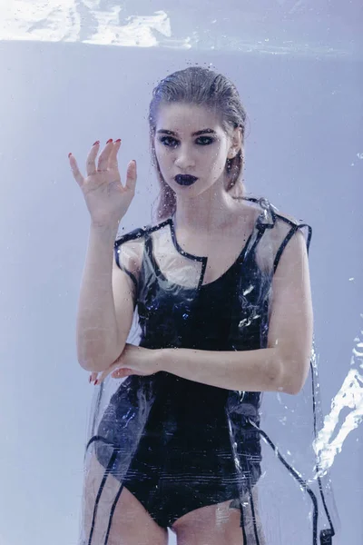Fashionable model with wet hair and dark lipstick dressed in black swimming suit and transparent rain coat is posing behind the wet  transparent membrane on the white background in the studio
