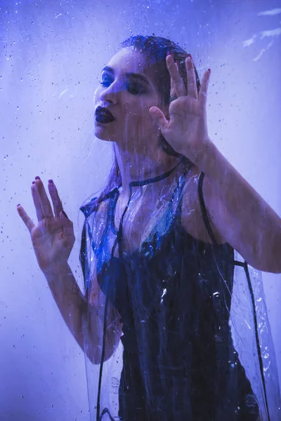 Fashionable model with wet hair dressed in black swimming suit and transparent rain coat is posing behind the wet transparent membrane in the studio with lighting simulating evening street light — Stock Photo, Image