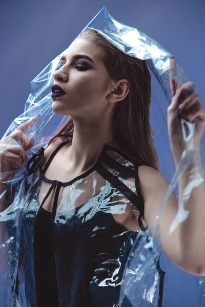Fashionable model with wet hair dressed in black swimming suit and transparent rain coat is posing under the transparent membrane in the studio with lighting simulating evening street light