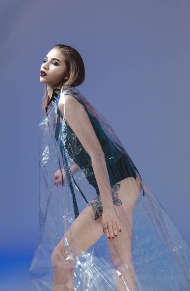 Beautiful young model with wet hair and tattoo on her leg dressed in black swimming suit and transparent rain coat is posing in the transparent membrane in the studio with lighting simulating evening