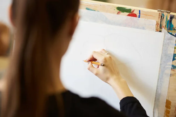 Menina bonita jovem de cabelos claros em óculos vestidos com blusa preta senta-se no cavalete e desenha um quadro com um lápis na escola de desenho — Fotografia de Stock