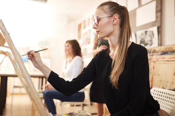 Niza joven en gafas vestidas con blusa negra y jeans se sienta en el caballete y pinta un cuadro en la escuela de dibujo — Foto de Stock