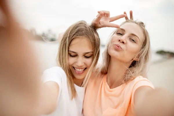 Dos chicas rubias jóvenes y atractivas se toman una selfie en la playa en un cálido día ventoso —  Fotos de Stock