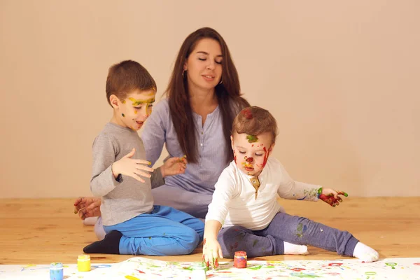 Jeune mère et ses deux petits fils avec les peintures sur leurs visages habillés en vêtements de maison sont assis sur le plancher de bois dans la pièce et la peinture avec les doigts sur le papier — Photo