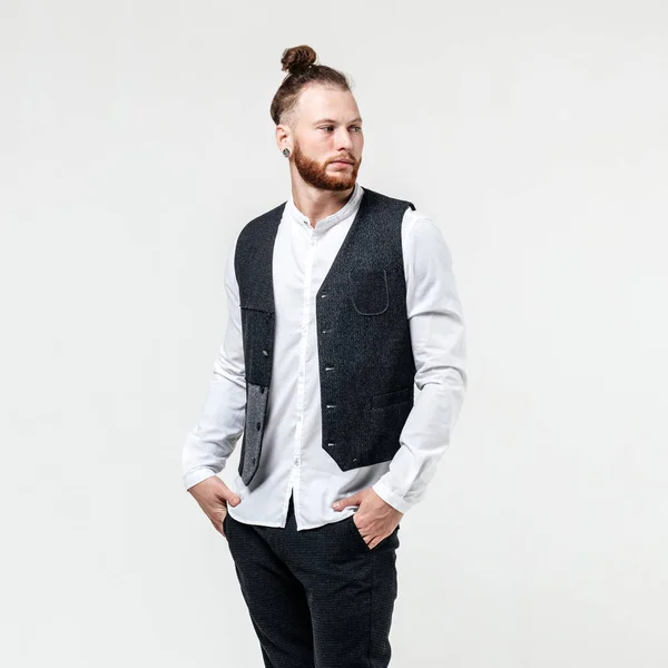 Bonito homem elegante com barba e penteado de pão vestido com colete cinza sobre uma camisa branca e calças cinza posa no estúdio no fundo branco — Fotografia de Stock