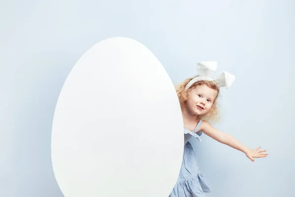 Маленькая кудрявая девочка в голубом платье с кроличьими ушами на голове смотрит из-за большого белого яйца на голубую стену. Пасхальный кролик — стоковое фото