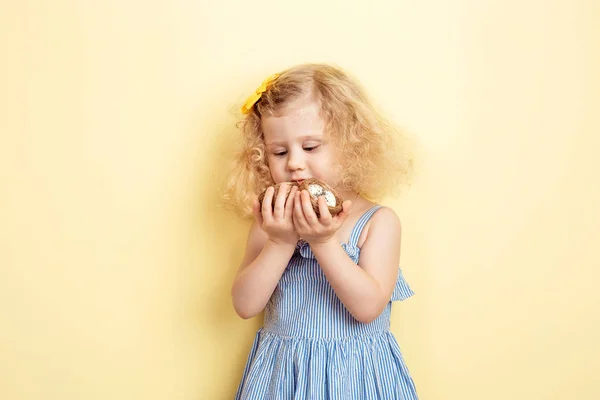 Kedves kis göndör lány a világoskék ruhát tart két kis fészket, tojást, a háttérben a sárga fal. — Stock Fotó