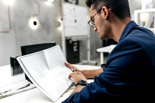 Elegante arquitecto de cabello oscuro en gafas y en una chaqueta azul está trabajando con documentos en el escritorio de la oficina — Foto de Stock