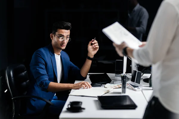 Arkitekten med briller kledd i blå jakke sitter ved pulten foran datamaskinen og ser og den andre mannen – stockfoto