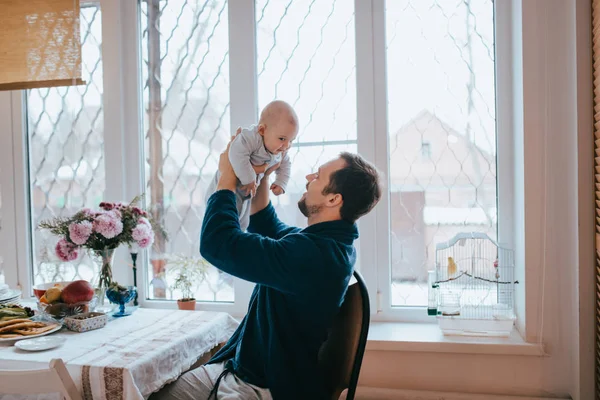 Ojciec trzyma w ramionach jego mały syn siedział na krześle obok duże okno w kuchni — Zdjęcie stockowe