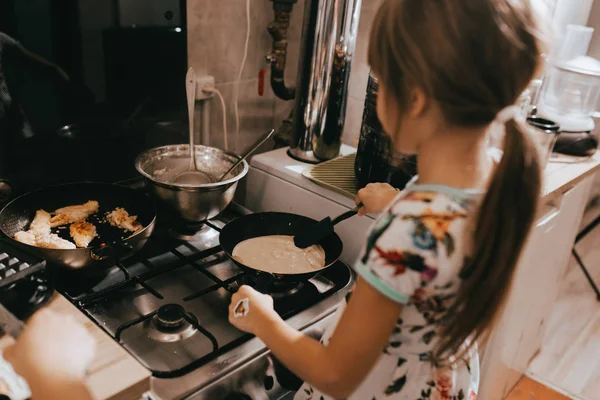 Menina ajuda sua mãe a cozinhar panquecas para o café da manhã na pequena cozinha acolhedora — Fotografia de Stock