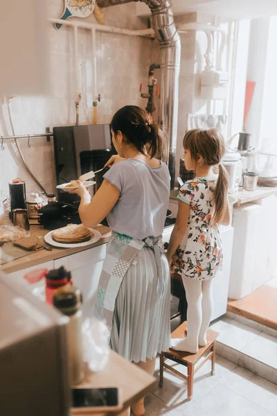 Little girl staying on the stool next to her mother is cooking pancakes for the breakfast in the little cozy kitchen — Stock Photo, Image