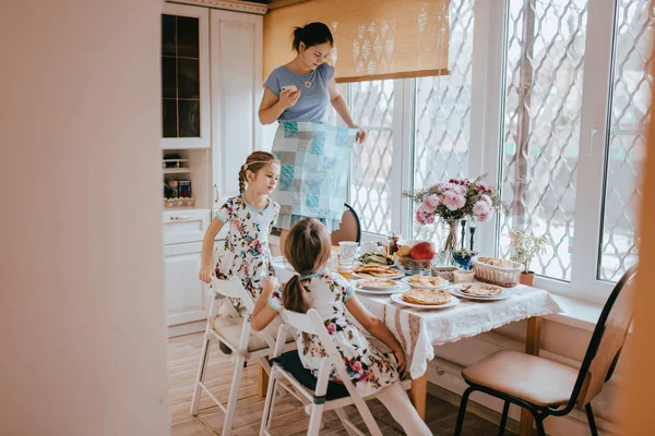 Mãe está de pé no banquinho e fazendo foto da mesa da cozinha com diferentes cursos para o café da manhã e sua filhinha sentada ao lado da mesa no banquinho — Fotografia de Stock