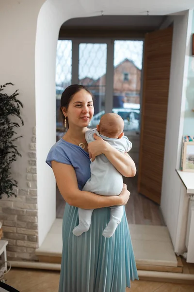 Młoda matka ubrana w lekki t-shirt niebieski i spódnica trzyma jej mały syn na ramionach w pokoju w domu — Zdjęcie stockowe