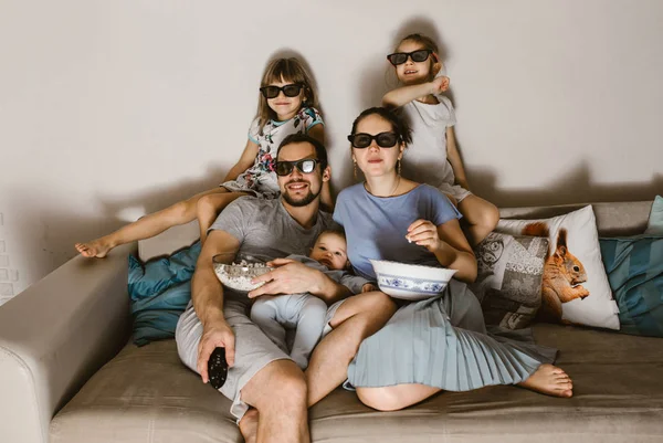 Όλα τα οικογενειακά πατέρα με το μωρό στην αγκαλιά του, τη μητέρα και δύο κόρες με τα ειδικά γυαλιά, βλέποντας τηλεόραση και τρώγοντας ποπ κορν που κάθεται στον καναπέ — Φωτογραφία Αρχείου