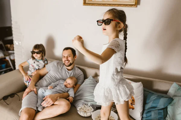Батько сидить на дивані з дитиною на руках і його дві дочки в спеціальних окулярах дивиться телевізор поруч з ним — стокове фото