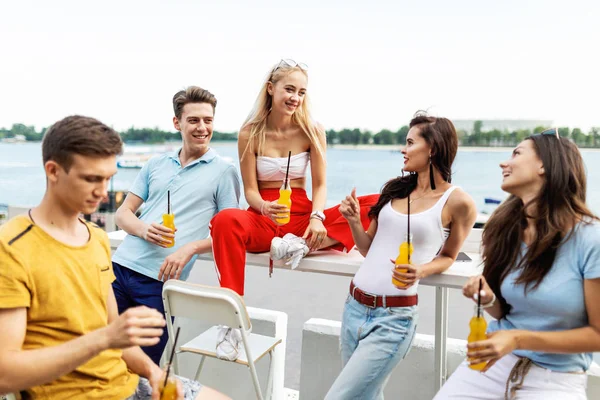 A company of good-looking friends laughing and drinking yellow cocktails in the nice cafe next to the river. Entertainment, having good time. River is in the background.