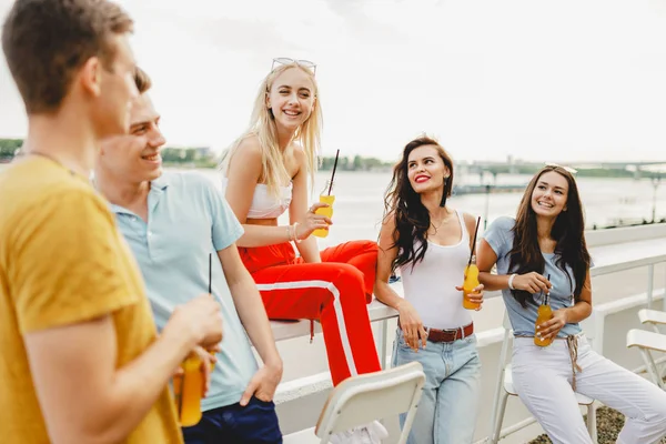 A company of good-looking friends laughing and drinking yellow cocktails in the nice cafe next to the river. Cheers. Entertainment, having good time. River is in the background.