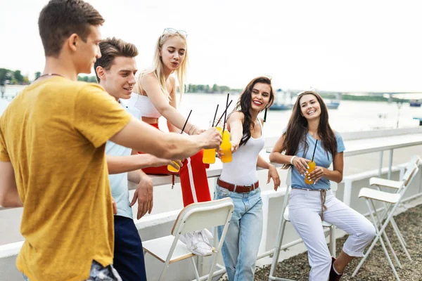 Uma companhia de amigos bonitos rindo e bebendo coquetéis amarelos no café agradável ao lado do rio. Um brinde. Entretenimento, divertindo-se. O rio está em segundo plano . — Fotografia de Stock