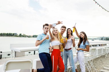 Gülüyor ve sarı kokteyl içme ve sosyalleşme ve selfie Nehri yanındaki güzel kafede yapma yakışıklı arkadaş şirket. Şerefe. Eğlence, iyi vakit geçiriyor. Nehir olduğunu