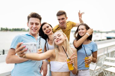 Gülüyor ve sarı kokteyl içme ve sosyalleşme ve selfie Nehri yanındaki güzel kafede yapma yakışıklı arkadaş şirket. Şerefe. Eğlence, iyi vakit geçiriyor. Nehir olduğunu