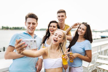 Gülüyor ve sarı kokteyl içme ve sosyalleşme ve selfie Nehri yanındaki güzel kafede yapma yakışıklı arkadaş şirket. Şerefe. Eğlence, iyi vakit geçiriyor. Nehir olduğunu