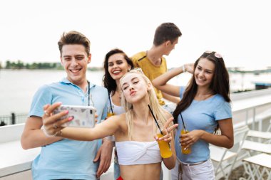 Gülüyor ve sarı kokteyl içme ve sosyalleşme ve selfie Nehri yanındaki güzel kafede yapma yakışıklı arkadaş şirket. Şerefe. Eğlence, iyi vakit geçiriyor. Nehir olduğunu