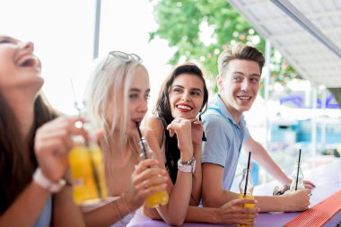 Gülüyor, sarı kokteyl içme ve güzel yaz kafe barda sosyalleşme yakışıklı arkadaş şirket. Arka planda nehirdir. Şerefe. Eğlence, iyi vakit geçiriyor