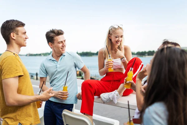 A company of good-looking friends laughing and drinking yellow cocktails and socialising in the nice cafe next to the river. Cheers. Entertainment, having good time. River is in the background.