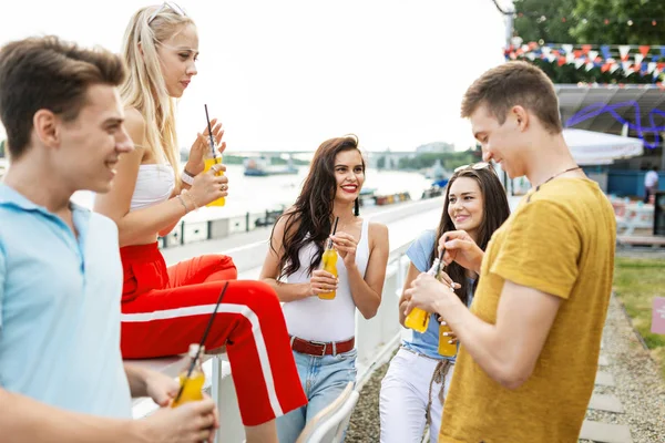 Una compañía de amigos guapos que ríen y beben cócteles amarillos en el agradable café junto al río. ¡Salud! Entretenimiento, pasándolo bien. Río está en el fondo . — Foto de Stock