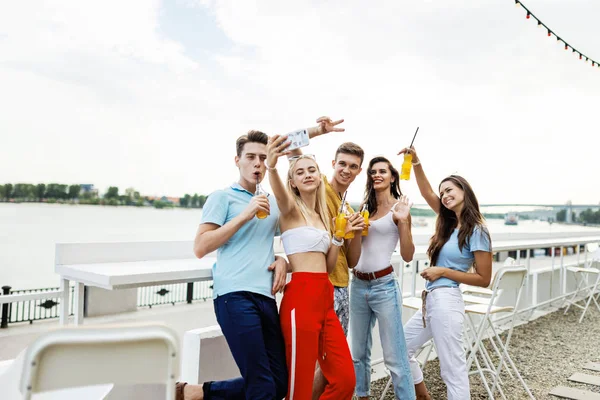 Una compañía de amigos guapos riendo y bebiendo cócteles amarillos y socializando y haciendo selfie en el agradable café al lado del río. ¡Salud! Entretenimiento, pasándolo bien. Río está en el — Foto de Stock