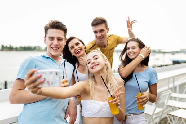 Una compañía de amigos guapos riendo y bebiendo cócteles amarillos y socializando y haciendo selfie en el agradable café al lado del río. ¡Salud! Entretenimiento, pasándolo bien. Río está en el —  Fotos de Stock