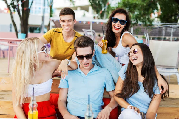 Una compañía de amigos guapos que usan gafas de sol riendo y bebiendo cócteles amarillos y socializándose en la mesa en el agradable café de verano. ¡Salud! Entretenimiento, pasándolo bien. Amistad . — Foto de Stock