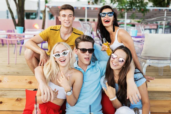 A company of good-looking friends wearing sunglasses laughing and drinking yellow cocktails and socialising at the table in the nice summer cafe. Cheers. Entertainment, having good time. Friendship.