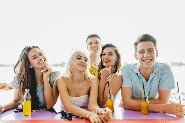 Uma companhia de amigos bonitos rindo, bebendo coquetéis amarelos e socializando no bar no agradável café de verão. O rio está no fundo. Um brinde. Entretenimento, se divertindo — Fotografia de Stock