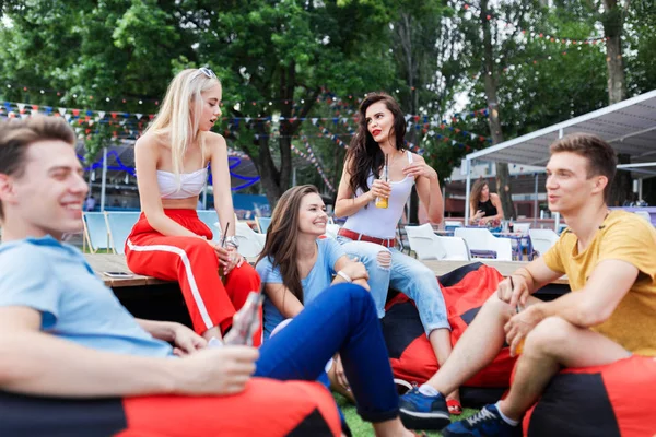 A company of good-looking friends laughing, drinking yellow cocktails and socialising while sitting on the bench and on the lawn in the nice summer cafe. Cheers. Entertainment, having good time