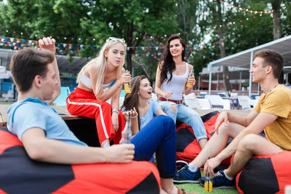 Uma companhia de amigos bonitos rindo, bebendo coquetéis amarelos e socializando enquanto se senta no banco e no gramado no agradável café de verão. Um brinde. Entretenimento, se divertindo — Fotografia de Stock