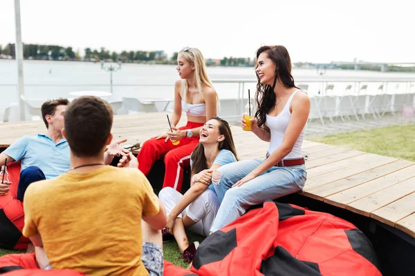 Une compagnie de beaux amis riant, buvant des cocktails jaunes et socialisant assis sur le banc et des sacs de haricots rouges sur la pelouse dans le joli café d'été. À la tienne. Divertissement, ayant — Photo