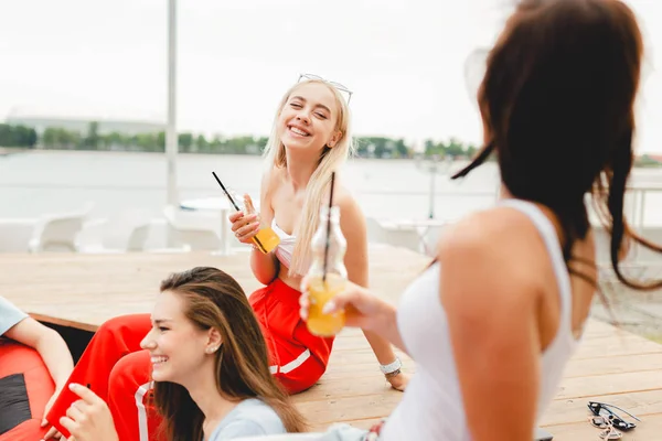 A company of good-looking friends laughing, drinking yellow cocktails and socialising while sitting on the bench and red bean-bags on the lawn in the nice summer cafe. Cheers. Entertainment, having