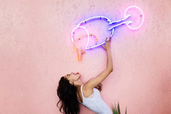 Das Mädchen posiert an der rosa Wand und gibt vor, aus einem Glas aus Neonröhren zu trinken, das an der Wand befestigt ist — Stockfoto