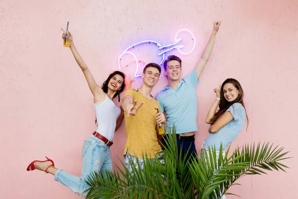 A company of good-looking friends laughing, drinking yellow cocktails is standing in front of the pink wall and behind a green plant in the nice summer cafe. Cheers. Entertainment, having good time