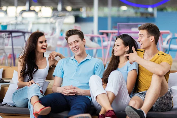 A company of good-looking friends is laughing and sitting on the bench with cushions in the nice summer cafe. Entertainment, having good time. Friendship.